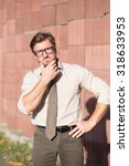 Small photo of Hipster businessman with look of verjuice touching his face outdoors. Handsome man in glasses keeping his hand on hip near red columns wall.