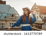 Young elegant fashionable  woman, model wearing trendy blue knitted sweater, wide leather belt, stylish white hat, earrings, posing at sunset, in European city. Copy, empty space for text