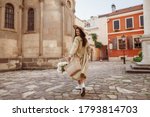 Happy smiling woman wearing straw hat, long trench coat, holding basket with flowers, walking in street of European city. Lifestyle, travel concept. Full-length portrait. Copy, empty space for text