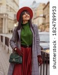 Small photo of Outdoor fashion, lifestyle portrait of happy smiling woman wearing trendy autumn outfit: orange hat, green turtleneck,midi coat, pleated skirt, wrist watch, with shoulder bag, walking in street