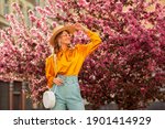 Happy smiling woman wearing trendy straw hat, yellow satin blouse, blue trousers, cat eye sunglasses, with wicker bag, posing in street near pink spring blossom trees. Copy, empty space for text 