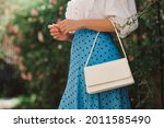 Summer street fashion details: close up of  classic white faux leather bag, small handbag in elegant outfit. Woman wearing trendy polka dot blue midi skirt,  posing in street. Copy, empty  space