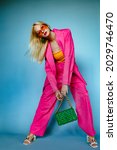 Small photo of Fashionable woman wearing trendy fuchsia color suit. orange sunglasses, holding stylish green quilted faux leather bag with chunky chain, posing on blue background. Full-length studio fashion portrait