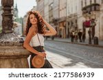 Beautiful happy smiling curly brunette woman wearing trendy summer outfit with round wicker shoulder bag, posing in street of European city. Fashion, lifestyle concept. Copy, empty space for text