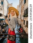 Small photo of Happy smiling fashionable blonde woman wearing straw hat, polka dot blouse, red skirt, holding patent leather handbag, posing on the bridge in Venice. Fashion, travel, lifestyle conception