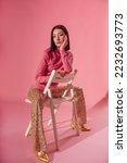 Small photo of Fashionable confident woman wearing trendy 70s style outfit with pink turtleneck top, sequined flare trousers, golden pointed toe shoes. Full-length studio portrait