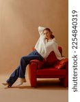 Small photo of Fashionable confident woman wearing trendy white knitted zip neck sweater, blue wide leg jeans, leather ankle boots, sitting on brown armchair, posing on beige backdrop. Full-length studio portrait