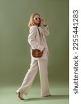 Small photo of Fashionable confident woman wearing elegant white suit with blazer, wide leg trousers, trendy sunglasses, brown suede shoulder bag, posing on green background. Full-length studio fashion portrait