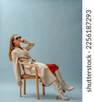 Small photo of Fashionable confident woman wearing elegant white woolen coat, sunglasses, high leather heeled boots, posing on blue background. Full-length studio fashion portrait. Copy, empty space for text