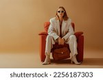 Fashionable confident woman wearing elegant white suit, sunglasses, ankle boots, holding classic brown leather bag, sitting in armchair, posing on beige background. Copy, empty space for text