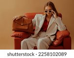Small photo of Fashionable confident woman wearing elegant white suit, sunglasses, chunky chain, holding classic brown leather bag, sitting in armchair, posing on beige background. Copy, empty space for text