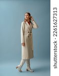 Small photo of Fashionable confident woman wearing elegant white woolen coat, high leather heeled boots, posing on blue background. Full-length studio fashion portrait. Copy, empty space for text