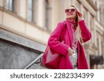 Fashionable happy smiling woman wearing fuchsia color coat, sunglasses, pink turtleneck, holding faux leather tote, shopper bag, posing in street of city. Copy, empty space for text