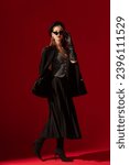 Small photo of Fashionable confident woman wearing black cat eye sunglasses, leather beret, gloves, waistcoat, boucle blazer, pleated midi skirt, high heel sock boots, posing on red background. Full-length portrait