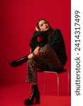 Small photo of Fashionable confident woman wearing trendy green turtleneck sweater, leopard print jeans, black leather platform chunky heel boots, posing on red background. Full-length studio fashion portrait