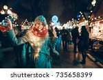 Outdoor photo of young beautiful happy smiling girl holding sparklers, posing in street. Festive Christmas fair on background. Model wearing stylish winter coat, knitted beanie hat, scarf.