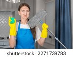 Small photo of Sad young woman in uniform looks hopefully at the camera holding a mop and sponge in her hands. Unwillingness to clean the apartment, fatigue after cleaning