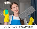 Small photo of Sad young woman in uniform looks at the camera with a misunderstanding, holding a mop and sponge in her hands. Unwillingness to clean the apartment, fatigue after cleaning.