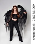 Small photo of Full length portrait of beautiful woman with long red hair wearing sheer corset top, leather pants, large black angel feather wings. Standing pose, walking forwards with gestural hands reaching out.
