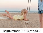 Small photo of Mother rides her little daughter on swing on the seashore. Cute towheaded girl on rope swing on the beach on sea background