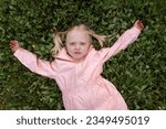 Small photo of Portrait of blonde girl with two ponytails in pink jacket lies on green grass. Towheaded small girl outside. Spring day.