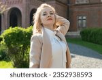 Small photo of Towheaded caucasian young woman walking in park on sunny warm spring day. Portrait of stylish girl in beige coat