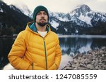 Young bearded man wearing yellow puff jacket and green hat standing near a mountain lake in winter. Snow mountain on background.  