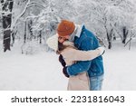 Young stylish loving couple kissing in winter park. Man and woman hugging while walking and enjoying snowy weather outdoors under falling snow. Valentine's day