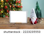 Horizontal tablet mockup on wooden table with Christmas decoration. Gnome and Christmas tree. Space to place text on the tablet screen.