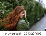 Small photo of Fashion woman in the city outside posing with a smile against buildings and trees with red long flying hair in the windy weather