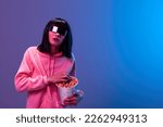 Shocked awesome brunet woman in pink hoodie trendy specular sunglasses eat popcorn open mouth posing isolated in blue violet color light background. Neon party Cinema concept. Copy space