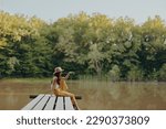 A young happy hippie woman sitting on the lake shore on a bridge in eco clothing made of natural materials in harmony with nature in the fall