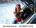 Happy woman holds a thermos and drinks tea sitting in car trunk in  winter forest. Rest, relaxation, travel, lifestyle concept.