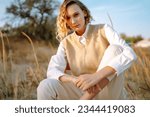 Small photo of Slender model with windy hair in stylish clothes poses among the wild grass on the beach. Beautiful woman standing on the windy coast. Freedom. Concept of fashion, relaxation, recreation.