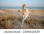 Small photo of Slender model with windy hair in stylish clothes poses among the wild grass on the beach. Beautiful woman standing on the windy coast. Freedom. Concept of fashion, relaxation, recreation.