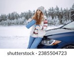 A smiling woman drinks a hot drink from a thermos while standing near her car on a winter snowy road in the forest. The concept of rest, freedom, relaxation, travel.