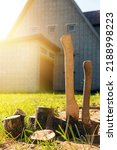 Small photo of Axes stand next to wooden firewood on the background of the house. Preparation of firewood for kindling. Stumps with axes, illuminated by sunlight. Chop wood. Axes next to sawn logs.