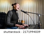 Small photo of Young Asian or Caucasian brunette woman having fun singing song on mic emotionally loud with pleasure and playing digital keyboard electronic piano in music studio or school, event or concert stage