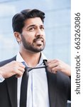 Small photo of Arabic serious angry irritated businessman or worker in black suit with beard standing in front of an office building and taking his tie off with his hands.