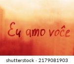 Handwritten message in Portuguese language Eu amo voce words I love you in english on misted glass on sunset window flooded with raindrops