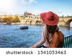 A traveler woman with a sunhat enjoys the view over the Vlatava River to the Charles Bridge and castle of Prague, Czech Republic, during a sunny autumn day