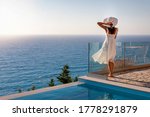 A elegant luxury woman in a white dress enjoys the summer sunset by the pool overlooking the Aegean Sea in Greece