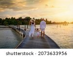 A family in white summer clothes walks holding hands over a wooden pier towards a tropical island in the Maldives during sunset time