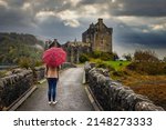 A tourist woman with a scottish pattern umbrella walks towards the Eilean Donan castle on a rainy autumn day, Scotland