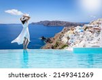 A beautiful woman in a white dress stands by the swimming pool and enjoys the view over the village of Oia, Santorini island, Greece, during her summer vacation time