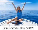 A happy, blonde woman with outstretched arms sits on a boat over calm, blue sea and enjoys her freedom