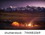 Night camping. Two men tourists sitting at the illuminated tent near campfire under amazing sunset evening sky in a mountains area. Snow mountain in the background