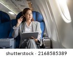 Small photo of Successful beautiful young asian business woman sits in airplane cabinplane and works on digital tablet with stylus. Flying at first class.