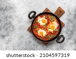 Small photo of Shakshuka in a frying pan on a gray rustic background. Poached eggs in a spicy tomato pepper sauce. Typical Jewish or Arabic food. Top view, flat lay. Textured object, selective focus.