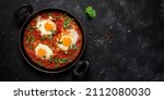 Small photo of Shakshuka eggs in a pan with toast on a black concrete background. Poached eggs in a spicy tomato pepper sauce. Traditional Jewish scrambled eggs. Top view, flat lay. Banner. Textured object, selectiv
